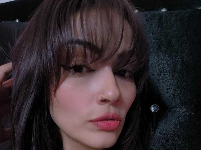 AliceGiorgia has dark brown hair, a nose piercing, and pink lipstick. She's looking at the camera with black eyeliner and her hand in her hair.