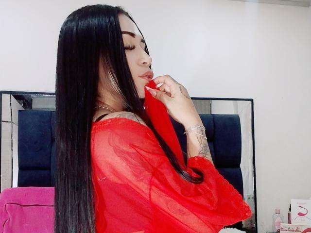 EvaMueller with long black hair, red sheer top, tattooed arm, holding red lipstick to her lips, eyes closed, looking sexy, standing in front of a dark blue headboard.