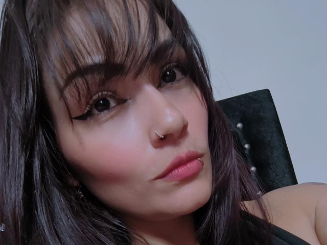 AliceGiorgia has dark brown hair, black eyeliner, a nose ring, and a lip ring, with pink lipstick and a slight pout, looking up with a serious expression.