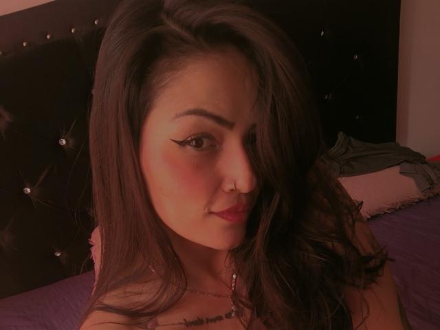 AliceGiorgia has long brown hair, pink lipstick, and a small nose ring. She's looking at the camera with a slight smile, wearing a pink top.