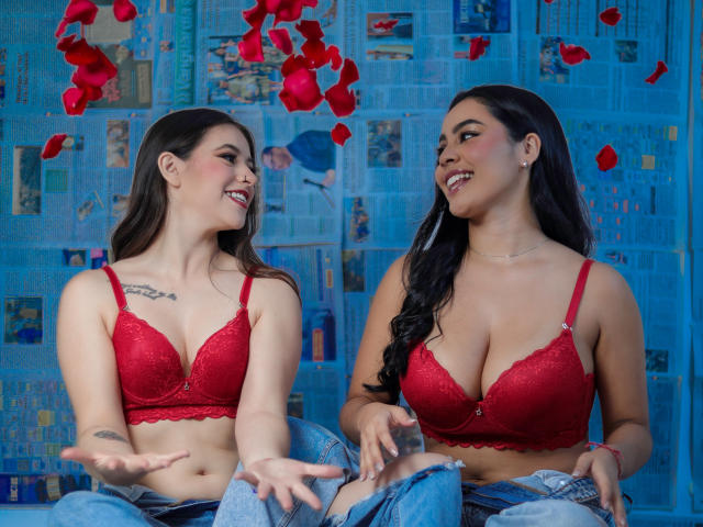 PaolaYKatty y PaolaYKatty con senos grandes en sostenes de encaje rojo, riendo y mirándose, sentadas con jeans, pétalos de rosa roja en el aire, ambas sonriendo..