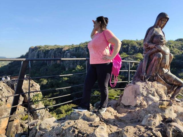 LadySelene with big tits, pink shirt, black pants, pink purse, standing next to bronze statue of seated woman, rocky overlook, metal railing, blue sky, green hills, daytime.