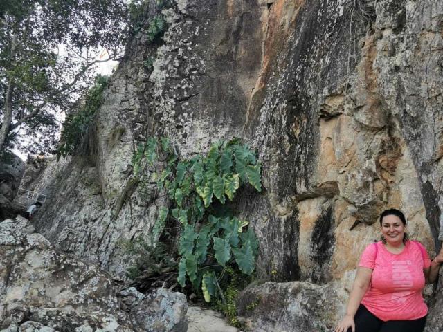 LadySelene with big tits, pink shirt, black pants, standing against a rocky cliff with green leaves. She's smiling, has dark hair, and is holding a rock.