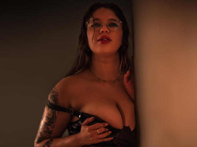 MissGaia with glasses, red lipstick, and black lace bra, showing her tits, tattoo on right arm, hand on chest, soft light, wall on right.