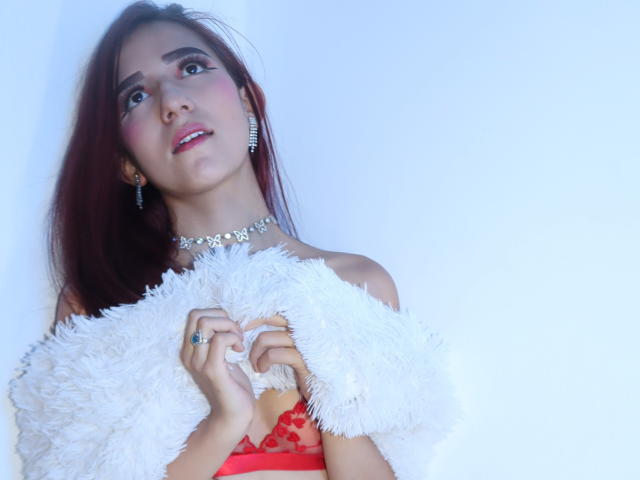NataliaPrada with red hair, pink blush, and red dress, holding a fluffy white off-shoulder, wearing a diamond choker and earrings, looking up with a soft smile.