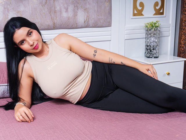 AnnieClarison, com longos cabelos pretos, deitada em uma cama rosa, vestindo uma regata bege e calças de ginástica pretas, mostrando tatuagens em seu braço direito, e sorrindo para a câmera..