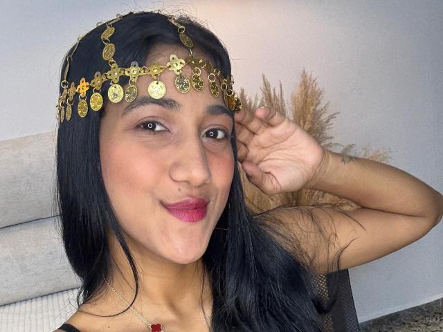 FfreyJa met zwart haar, die een gouden munt als hoofdtooi draagt, met een glimlach en roze lippen, de rechterhand op het hoofd, bruine ogen, een bruine huid en een rode ketting..