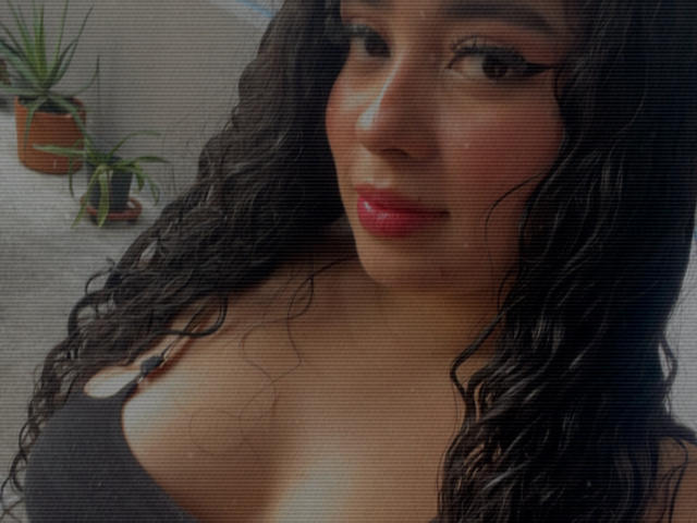 AgathaLynx with curly black hair, wearing a black tank top, showing her cleavage, pink lipstick, and a subtle smile. Her brown eyes are slightly open.