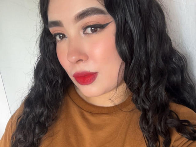 AgathaLynx has wavy black hair, red lipstick, and thick black eyeliner with a cat-eye look, wearing a brown turtleneck. Her face is pale with a slight blush.