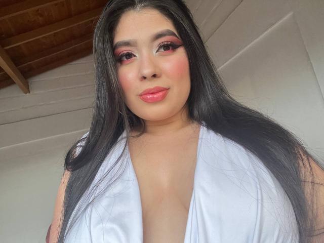 AgathaLynx with long black hair, pink eyeshadow, blush, and pink lipstick, wearing a low-cut white top, showing her big tits. Wooden ceiling in the background.