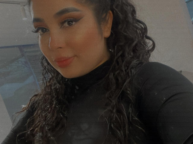 AgathaLynx has curly black hair, dark eyebrows, and is wearing a black turtleneck. She has red lipstick and is smiling slightly. Her tits are not visible.