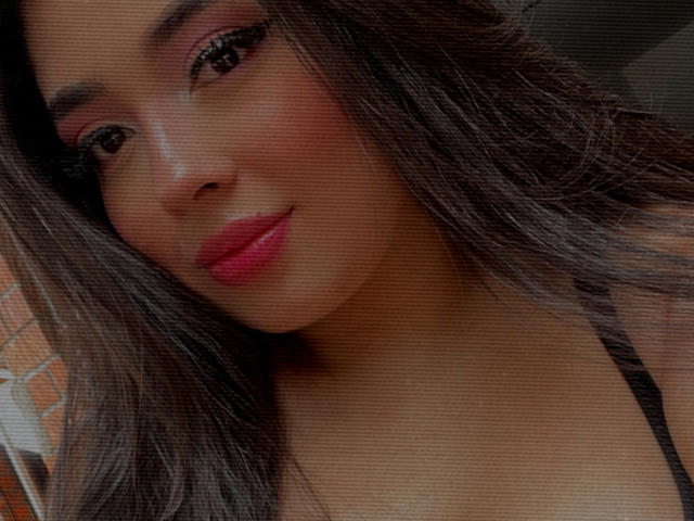 AgathaLynx has glossy pink lips, dark brown eyes, and long, wavy black hair. Her tan skin is smooth with a subtle glow. She's wearing a black top.