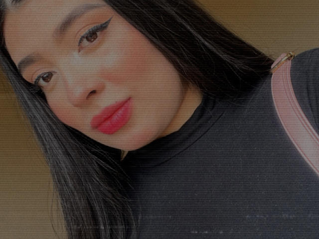 AgathaLynx has long black hair, red lipstick, and winged eyeliner. She's wearing a black shirt with a pink strap over her shoulder. Her face is slightly tilted.