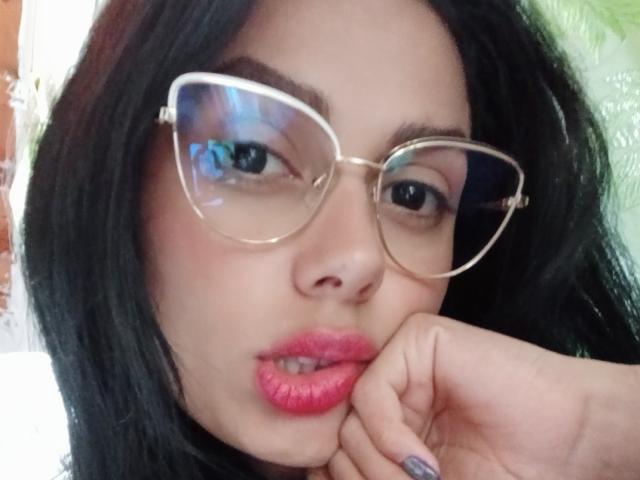 AnaisHilton with black hair, pink lips, and clear glasses, resting her chin on her hand, showing off her pink lipstick and a silver nail.