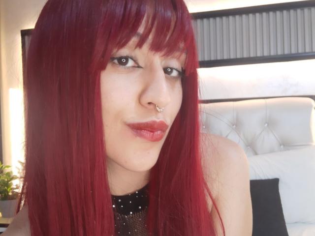 KaylinKrof has red hair, a nose ring, and red lipstick. She's smiling slightly with her red lips and wearing a black, sparkly top. Her eyes are dark and lined.