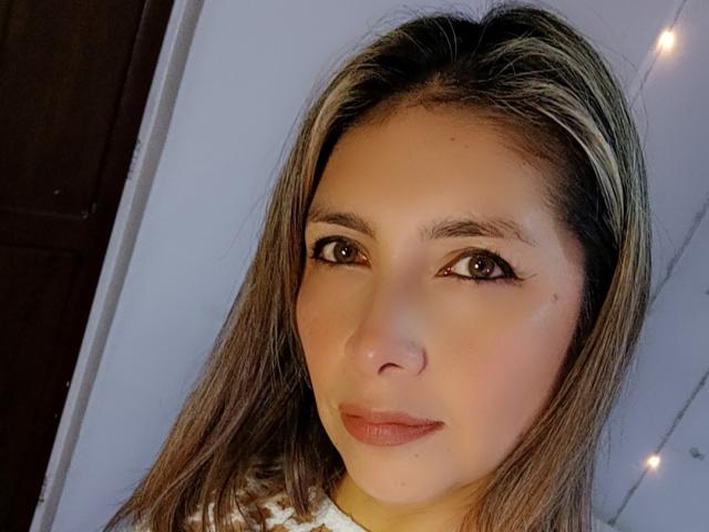 Het gezicht van Michelle Dumont, met bruine ogen, steil bruin haar en roze lippenstift. Ze draagt een witte top, en er zijn kleine lichtjes in de achtergrond..