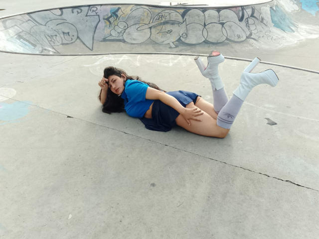 MargaritaDudole lying on concrete, wearing blue shirt, blue skirt, white knee socks, white heels, exposing her bare ass, with long dark hair, in a graffiti-covered skatepark.
