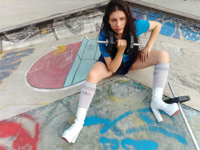 MargaritaDudole, squatting, blue shirt, black bra, white boots, gray leg warmers, holding dumbbell, concrete skatepark, long dark hair, serious expression, visible pussy through shorts, graffiti background.