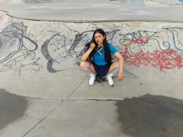 MargaritaDudole, squatting with her ass down, wearing a blue shirt, dark blue skirt, and white sneakers, in a graffiti-covered concrete bowl. Long black hair, hand on neck.