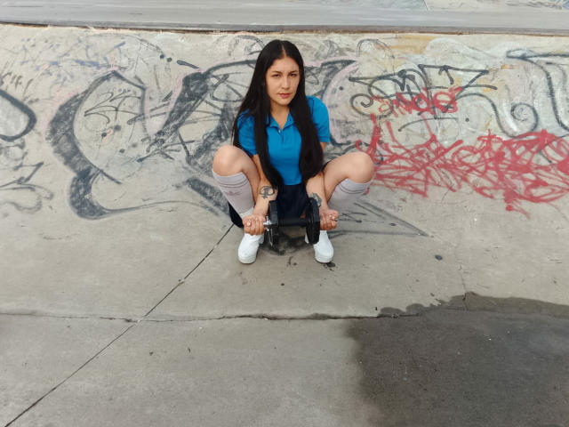 MargaritaDudole, squatting with knees apart, wearing a blue shirt, black skirt, gray socks, and white shoes, has long black hair, small tits, and a tattoo on her right wrist.