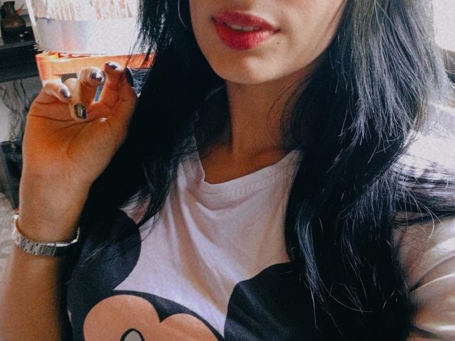 AnaisHilton with long black hair, red lipstick, white and black tee, showing hand with metallic nail polish, wearing silver watch, partial face visible.