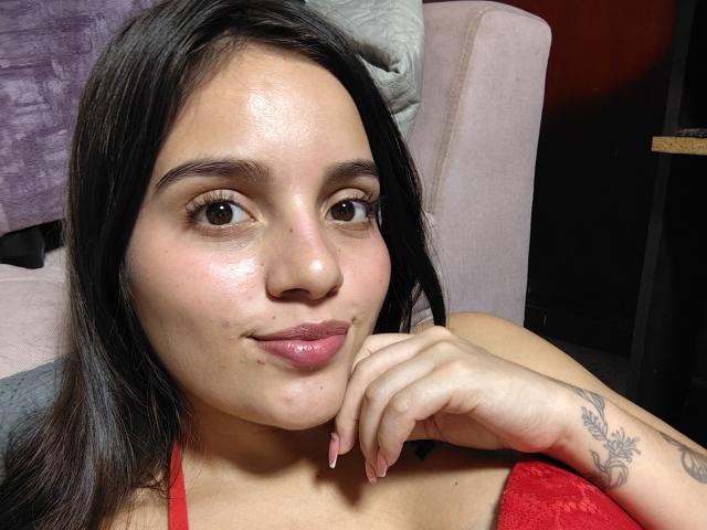 ShiaraSexy has dark hair, brown eyes, and glossy pink lips. She rests her chin on her hand, showing a tattoo on her right arm, in a red top.