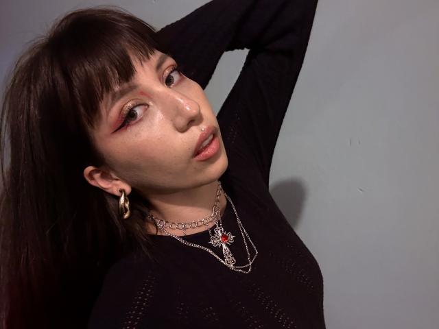 IzzysMisss with dark brown hair, raised arm, black sweater, gold hoop earrings, chain necklaces, pink eyeliner, nose ring, and slightly parted pink lips, looking up.