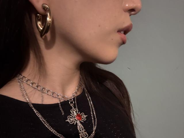 IzzysMisss's face, showing her nose and slightly open mouth. She's wearing a black shirt, silver chain necklaces with a cross and red gem, and large gold hoop earrings.
