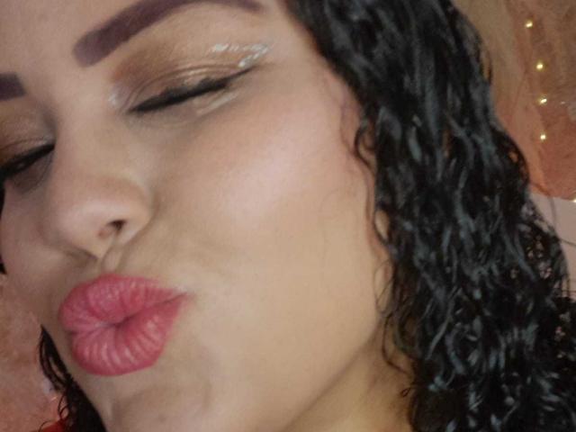 MelannieCarter's face with closed eyes, dark curly hair, and shiny pink lipstick, puckering her lips. She has well-defined eyebrows and subtle eye makeup. Her skin is tan.