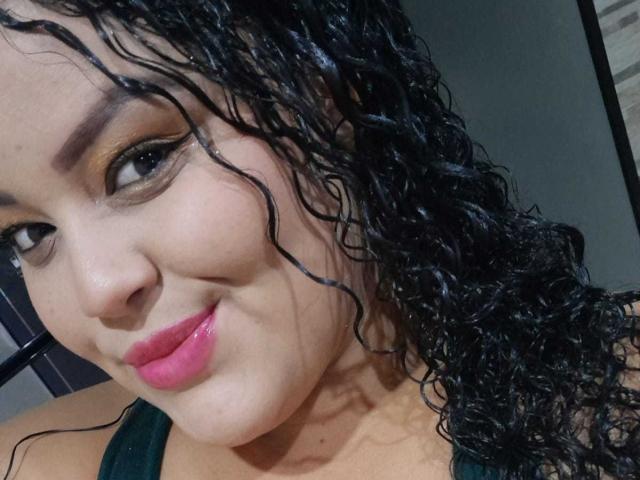 MelannieCarter has curly black wet hair, brown eyes, and pink lipstick. She's smiling with a slight tilt to her head, wearing a green top. Her ass is not visible.