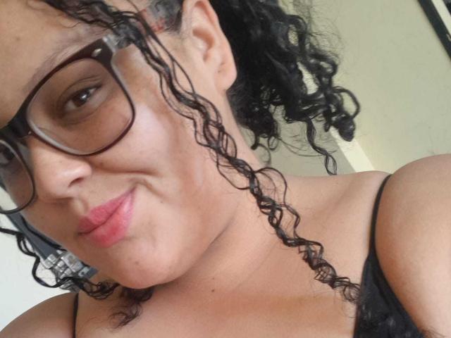 MelannieCarter with curly black hair, glasses, pink lipstick, and a slight smile, wearing a black spaghetti strap top, showing her boobs. She has a light brown skin tone.