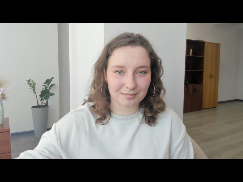Klarisia with curly brown hair, blue eyes, and small nose, wearing a white sweatshirt, large hoop earrings, and a slight smile, in a bright room.