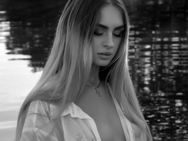 Black-and-white photo of PlayfulfBoo with long, straight blonde hair, looking down, wearing an open shirt, showing cleavage, a cross necklace, and soft, sexy eyes.