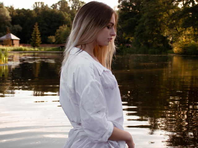 PlayfulfBoo with long blonde hair, white shirt, standing in water, looking to the side, showing her small tits and slim ass. Water reflects her and trees in background.