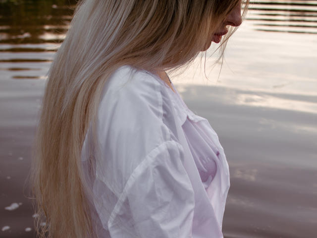 PlayfulfBoo with long blonde hair, white shirt, and visible small tits, standing by a water body, side profile, looking down.