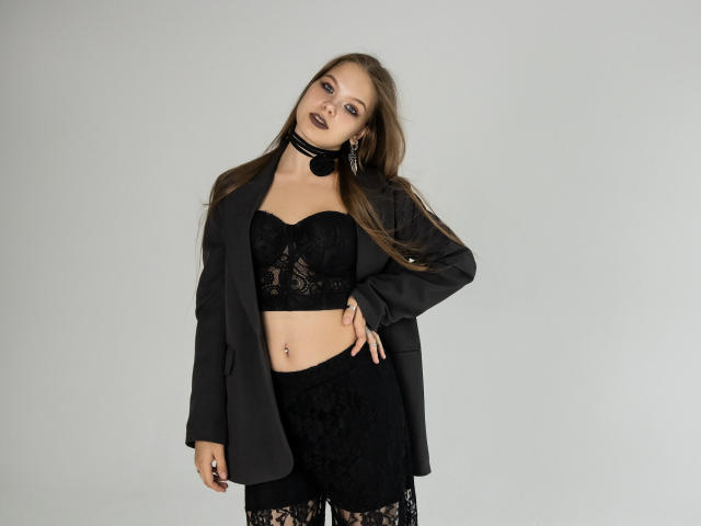 BethCutie con cabello largo y castaño, un sujetador de encaje negro, una chaqueta negra, shorts de encaje negro, un collar y lápiz labial negro, mostrando un piercing en el ombligo, de pie con confianza..