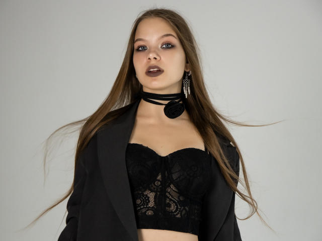 BethCutie, com cabelos castanhos longos, vestindo um corpete de renda preto, um blazer preto, um colar e batom escuro, olhando diretamente para a câmera, com a boca ligeiramente aberta..