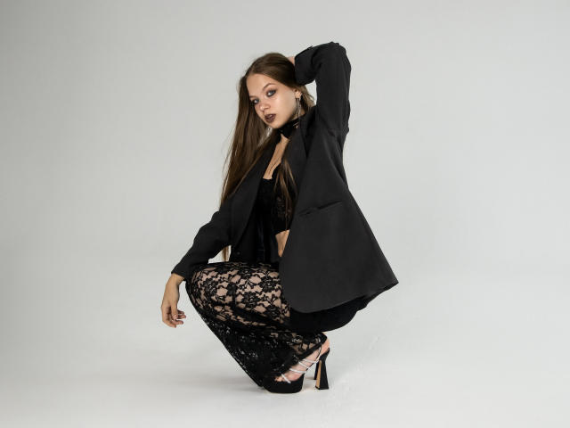 BethCutie, avec de longs cheveux bruns, accroupie, portant un pantalon en dentelle noire, un blazer noir, un soutien-gorge noir et des talons hauts, la main derrière la tête, avec un rouge à lèvres foncé, regardant l'appareil photo..