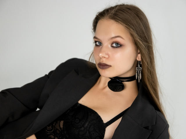 BethCutie met blauwe ogen, bruin haar, een zwarte kanten bh, een zwart blazer, een zwarte choker, donkere lipstick en oorbellen die bungelen, die verleidelijk naar de camera kijkt..