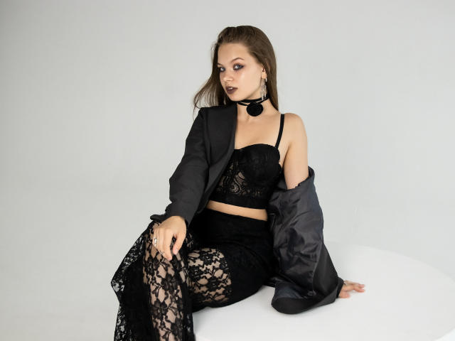 BethCutie, avec ses cheveux bruns, son soutien-gorge en dentelle noire, sa jupe assortie et sa veste grise ample, est assise sur du blanc. Elle porte un collier noir et des collants en dentelle, et a un air séduisant..