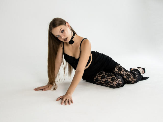 BethCutie mit langen braunen Haaren, einem schwarzen Spitzenoberteil und passenden Spitzenhosen, im Vierfüßlerstand, die in die Kamera schaut, mit schwarzem Choker, schwarzem Lippenstift und schwarzen High Heels..