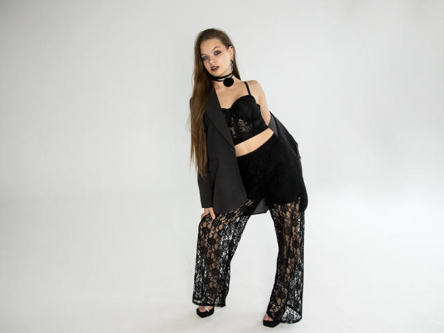 BethCutie with long brown hair, wearing black lace bra, black high-waist pants, black blazer, black choker, and open-toe heels, standing with a confident, sexy pose.