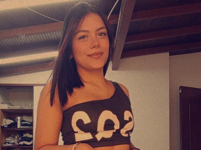 EmilyRoberts with medium brown tits in a black crop top with white letters, smiling slightly, brown hair, ceiling with ducts in background, light skin, subtle makeup.