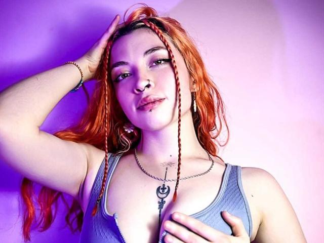 SharickAddler with red braids, nose ring, and lip piercing, wearing a gray tank top, hand on head, other hand on tits, purple-blue light, chain necklace with crescent moon.