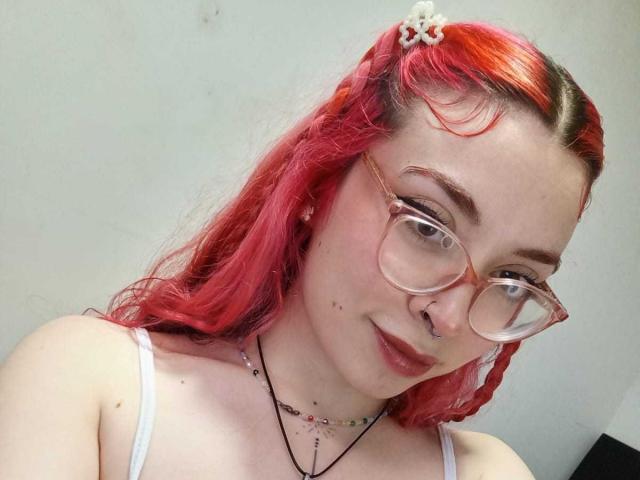 SharickAddler with red hair, glasses, nose ring, white top, and a small white hair clip, showing off her tits and a slight smile.
