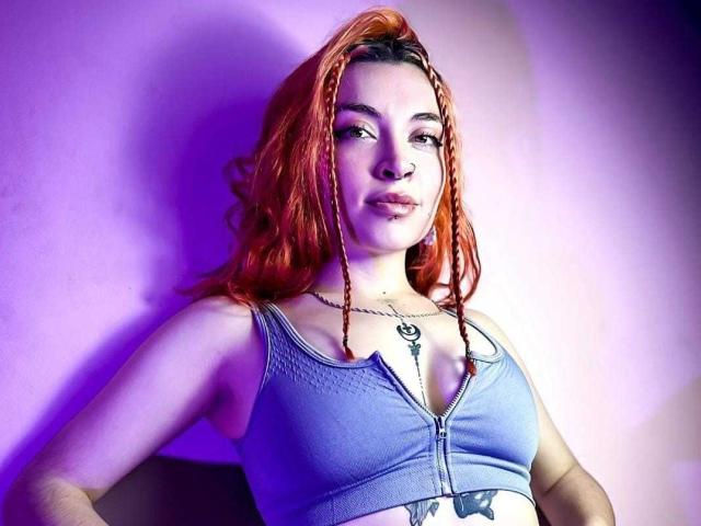 SharickAddler with red braids, blue zip-up top, showing tits, tattoo on belly, chain necklace, purple lighting, slightly smug expression, standing against pink wall.