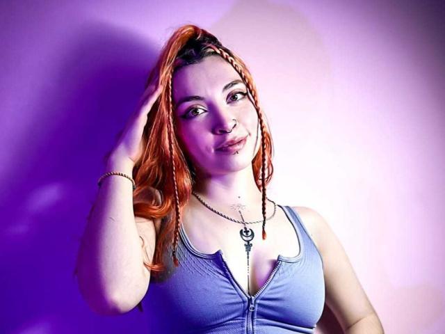 SharickAddler with orange braids, blue tank top, hand in hair, smiling, necklace, purple lighting, pale skin, small tits, visible nipple, subtle makeup, relaxed, confident, upper body shot.