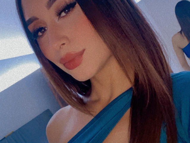 ValeriaKlaus with long brown hair, wearing a teal top, showing off her tits with an off-shoulder look, and red lips, looking sexy in the selfie.