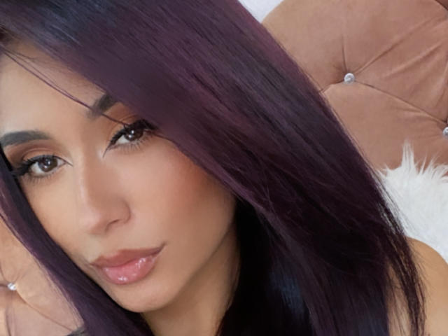 ValeriaKlaus tem cabelos longos e roxos escuros, sobrancelhas grossas, olhos castanhos, lábios rosados e brilhantes e uma pele bronzeada. Ela está usando um encosto de cabeça rosa e felpudo ao fundo..