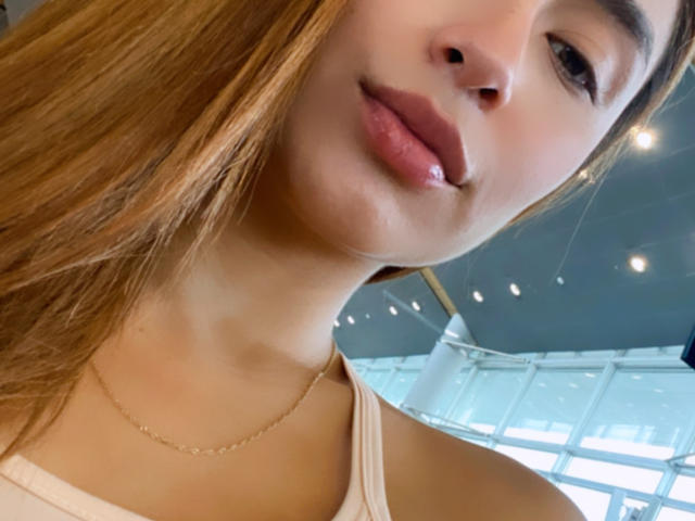 ValeriaKlaus's face, showing her glossy pink lips and light brown hair, wearing a thin gold necklace and a beige top, with a subtle smile and soft skin.